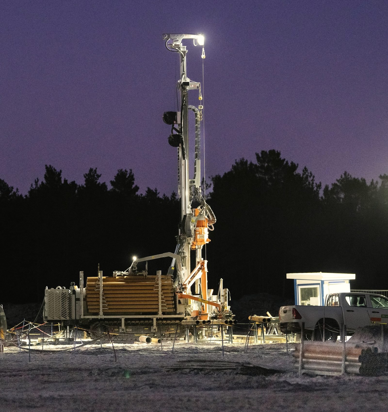 A drilling mast lit by work lights against a violet dusk sky, pipe laid out on the ground below